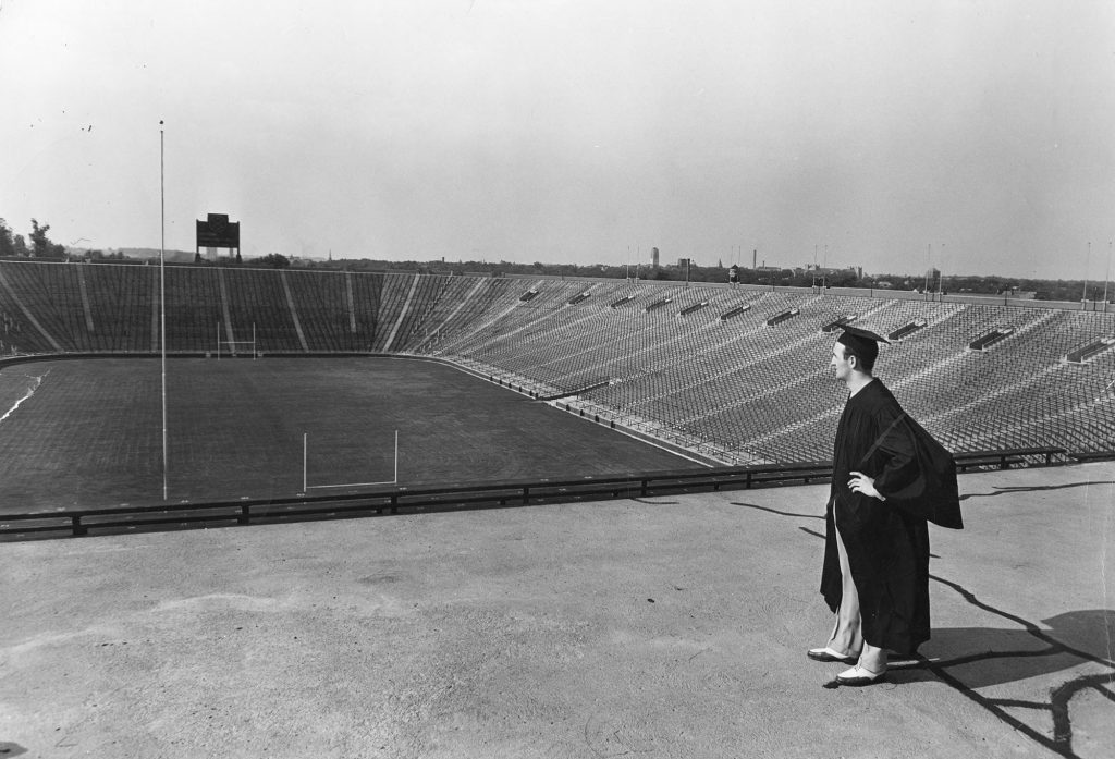 A grayscale photograph, circa 1941, of Tom Harmon in a graduation robe looking in to Michigan Stadium
